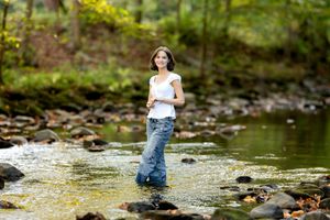 Young woman in white top and jeans standing in a rocky creek surrounded by lush green forest.