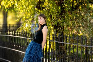Young woman in a blue skirt posing by an iron fence under golden sunlit tree branches.