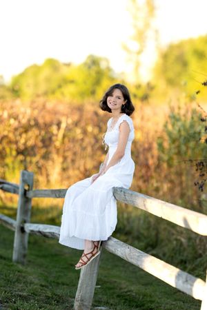 Smiling young woman in white dress sitting on rustic wooden fence in golden sunset light.