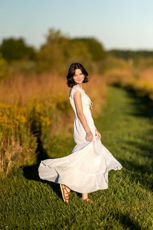 Young woman in flowing white dress twirling on a green path through a sun-drenched meadow.