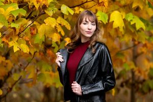 Young woman in leather jacket standing among vibrant yellow and orange autumn maple leaves.