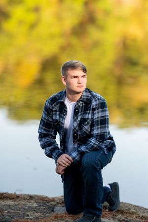 Teen boy in blue plaid flannel shirt kneels on rocks near a lake with fall foliage background.