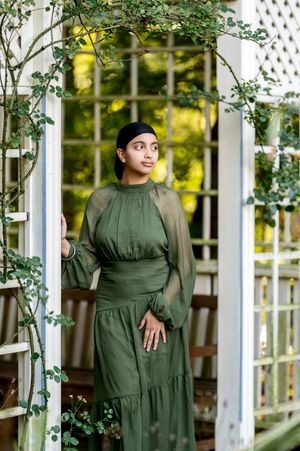Young woman in green maxi dress poses in white lattice garden structure with climbing vines.