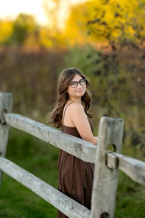 Teenage girl with glasses in a brown dress leaning on a rustic wooden fence at golden hour.
