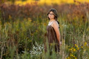 Teenage girl with glasses in a brown dress standing in a wildflower meadow during sunset.
