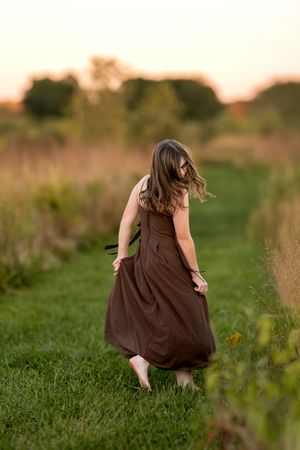 Young woman in a flowing brown dress walking barefoot through a green meadow path at sunset.