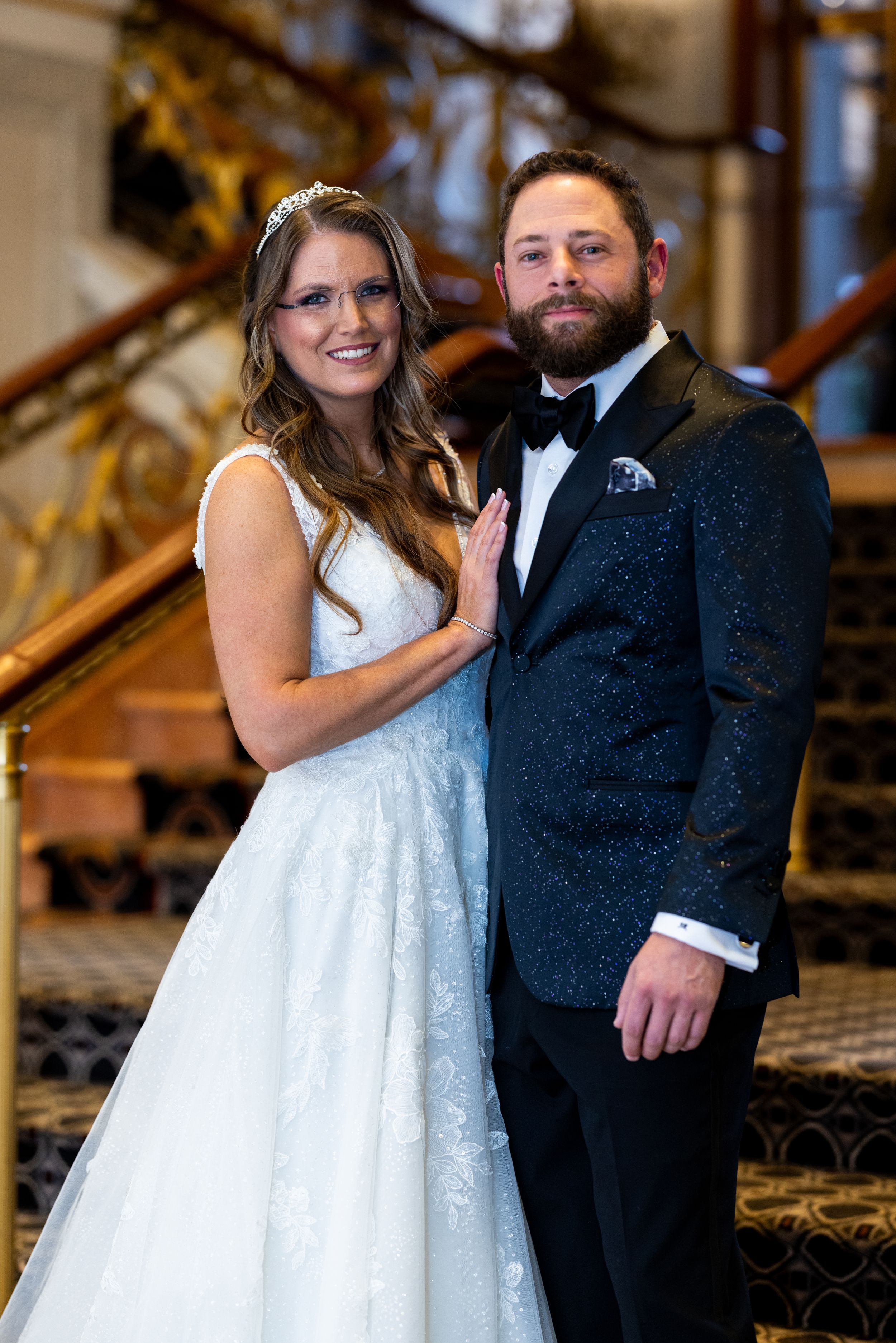 Newlyweds pose on elegant staircase with bride in lace gown and groom in sparkly tuxedo.