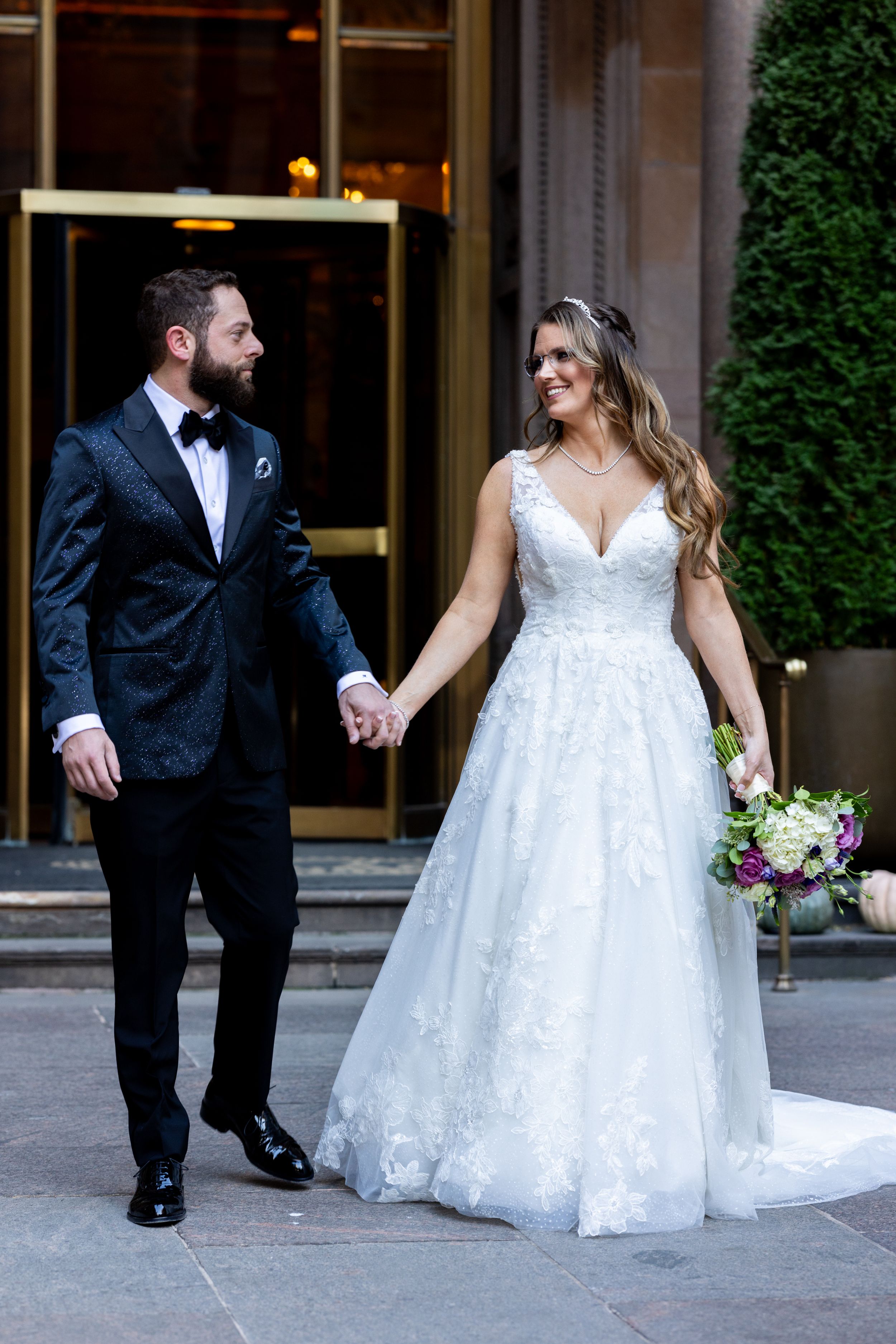 Couple in wedding attire walking outside luxury venue with beautiful landscaping.
