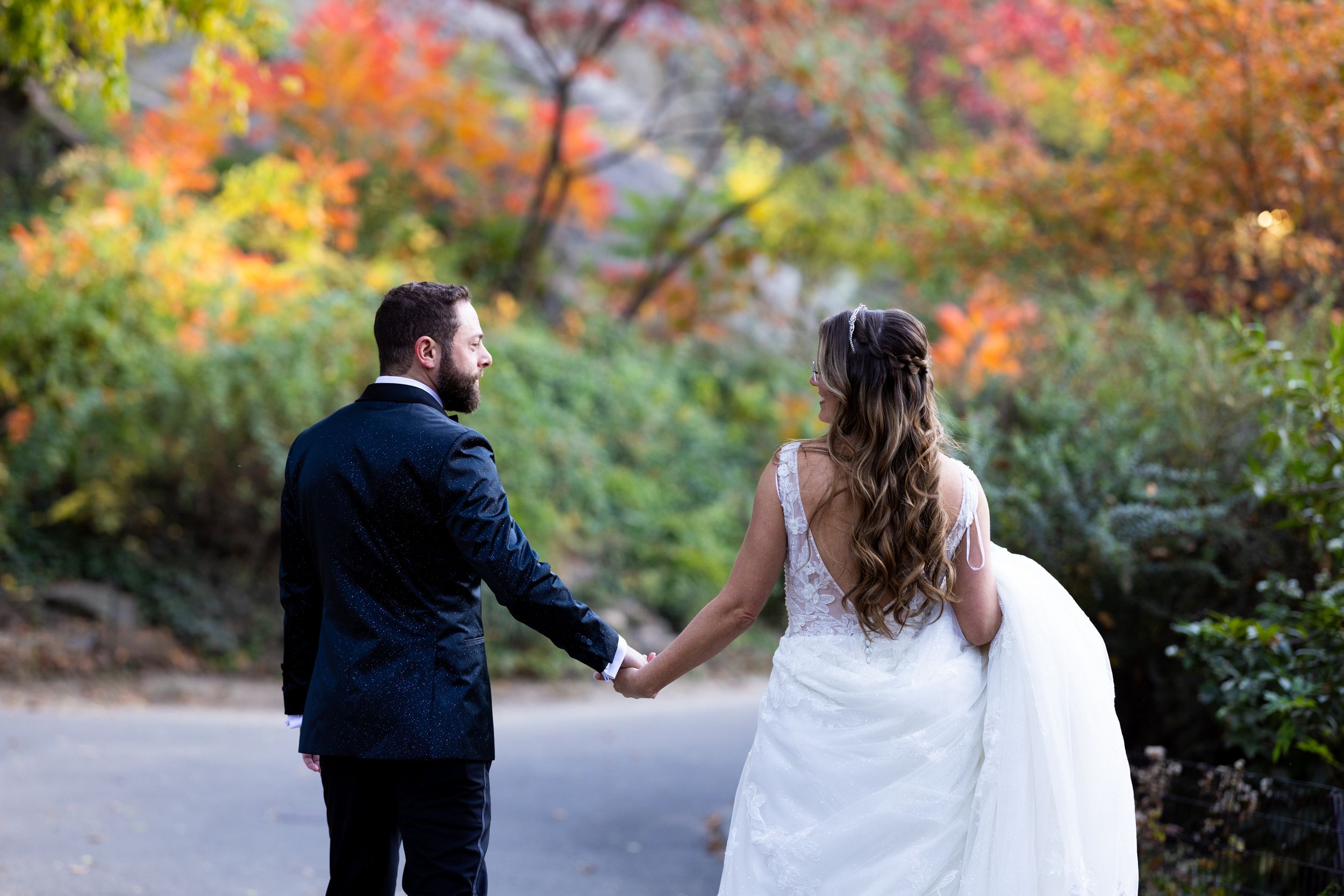 Couple holds hands walking through park with vibrant fall foliage.