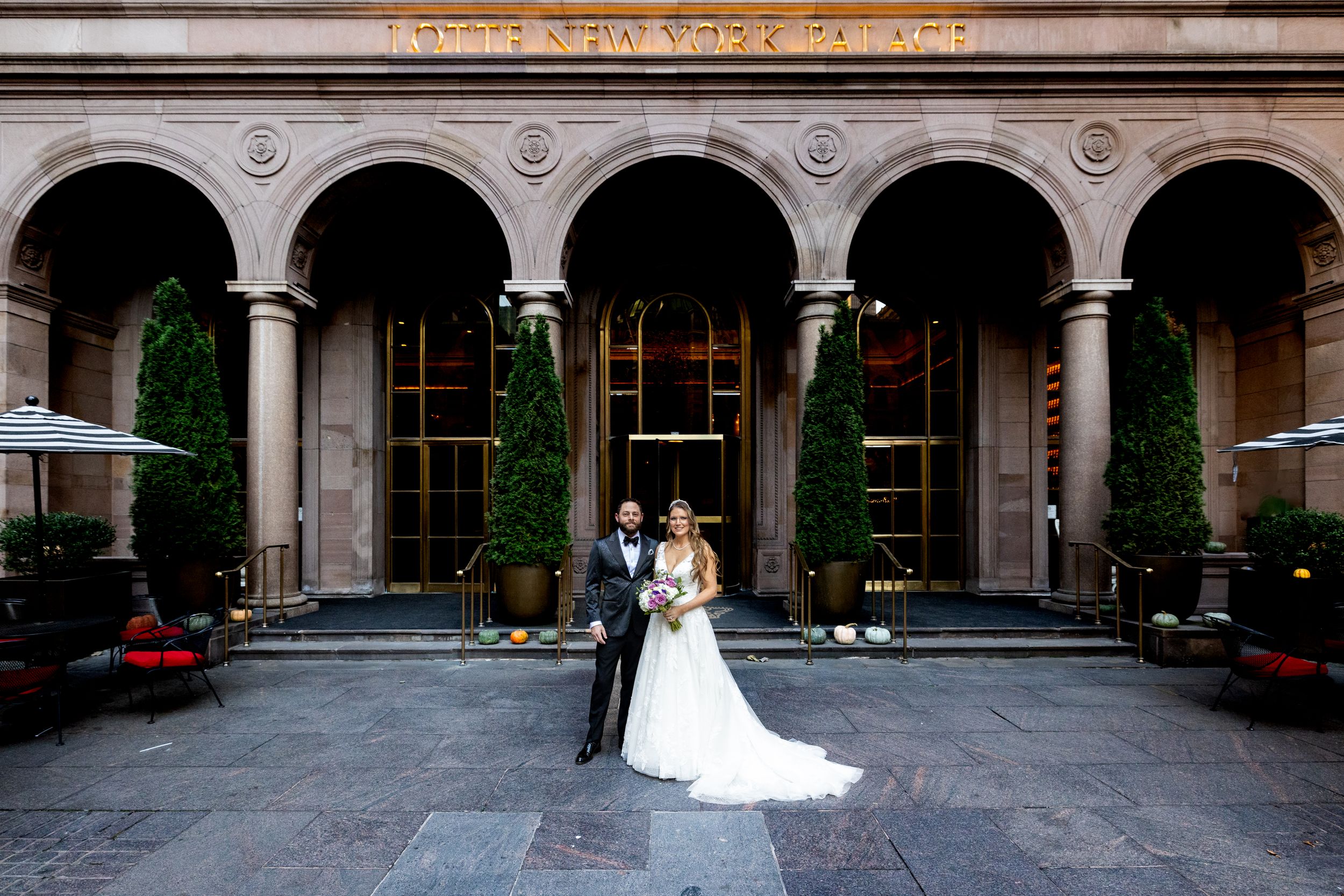 Wedding couple stands under beautiful stone archway at The Lotte New York Palace entrance with classic architecture.