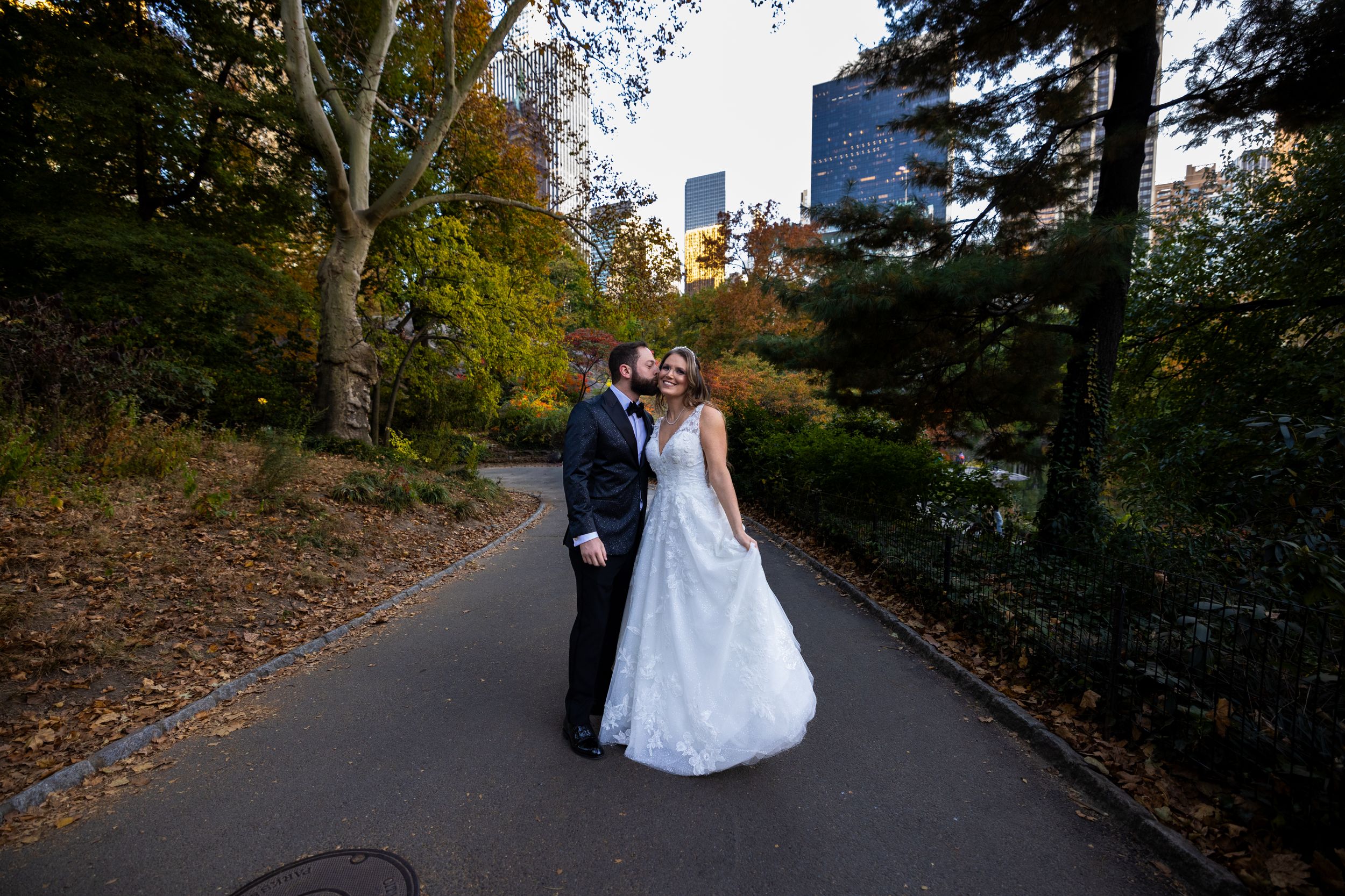 Wedding couple walking along autumn path with city skyline in background.