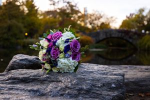 A vibrant purple and white wedding bouquet rests on a rocky surface with a blurred garden backdrop.