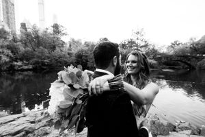 A romantic black and white wedding photo captured by a pond with city buildings in the background.