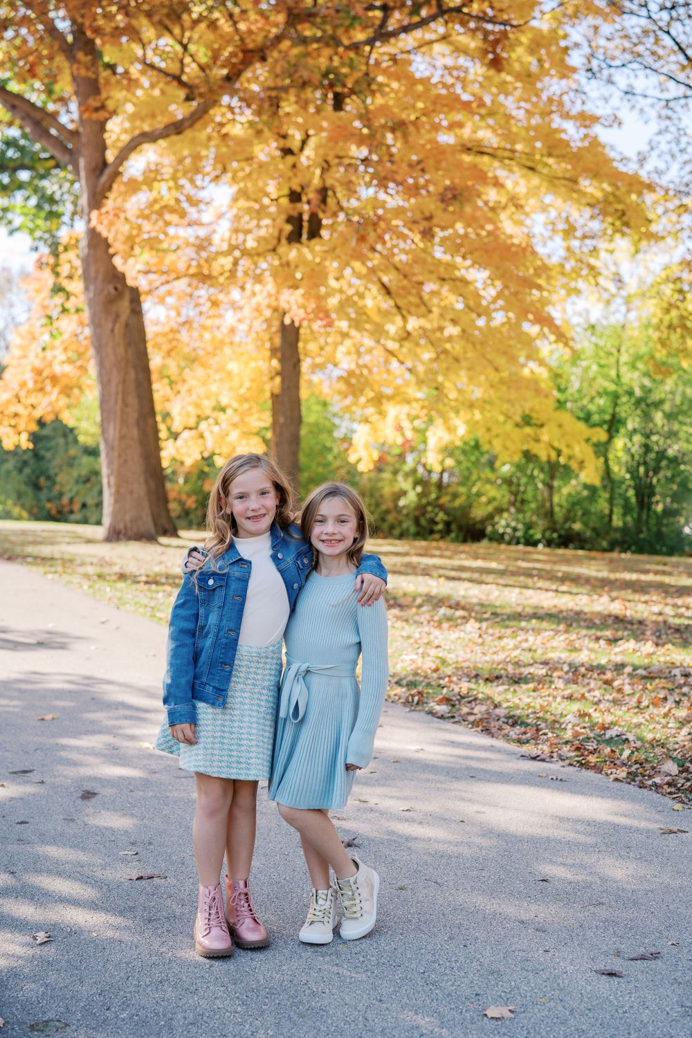 Two children in light blue outfits pose together under golden autumn trees.