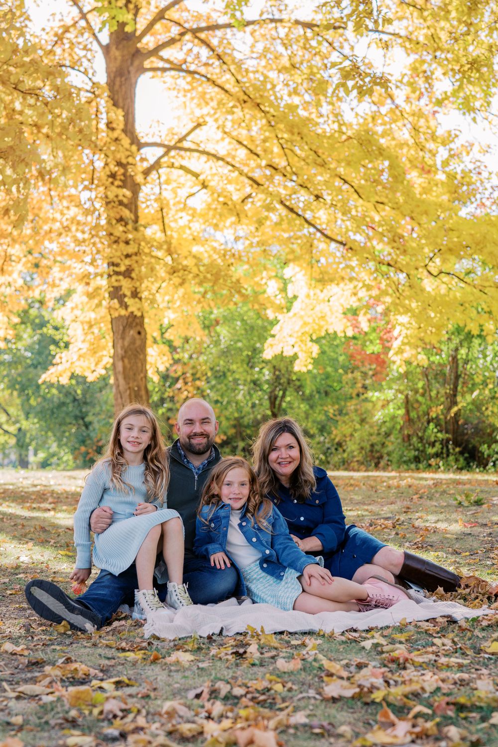Family lying on a blanket under golden fall trees during an outdoor photo session.