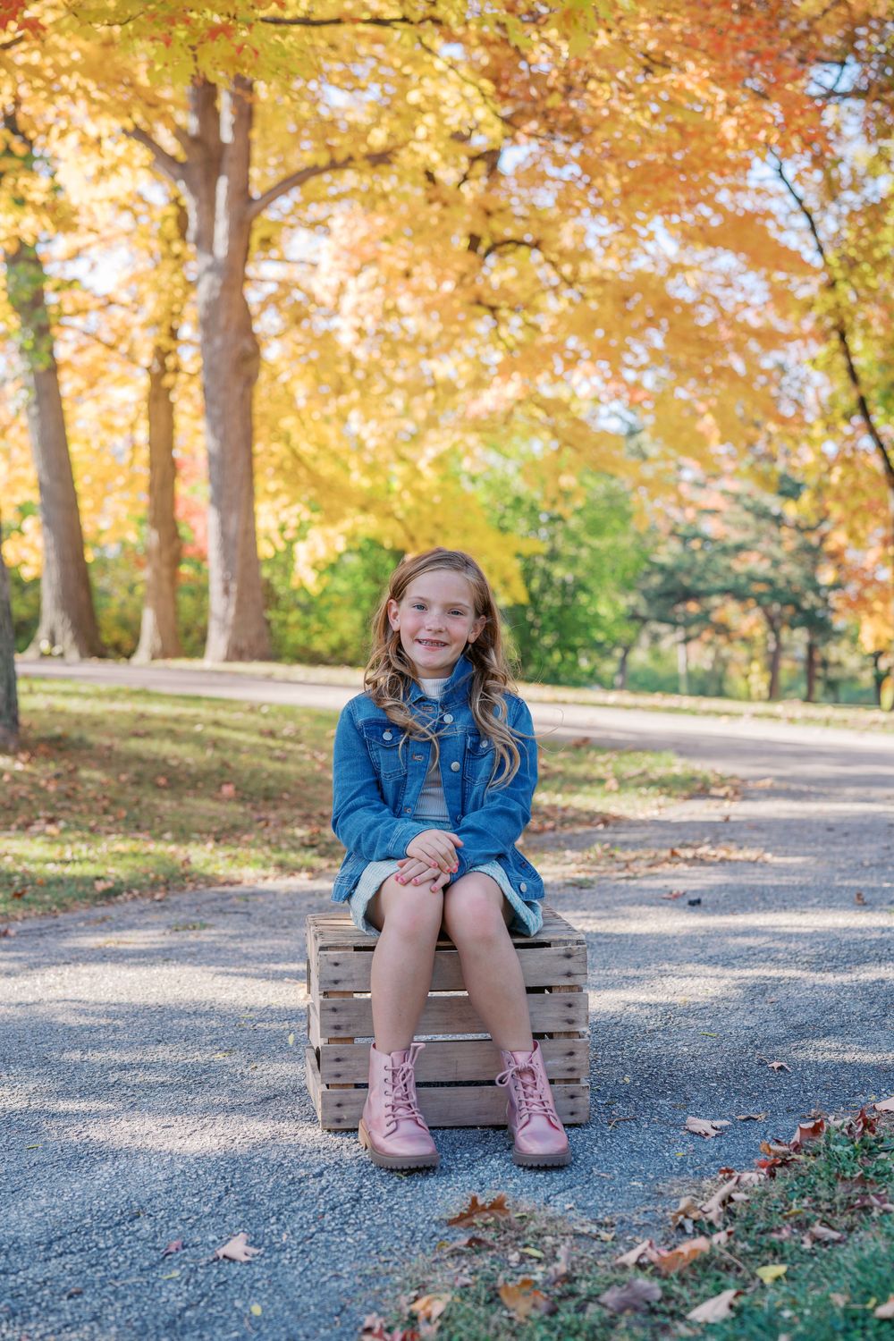 Young person sitting on wooden crate outdoors during autumn with trees in background.