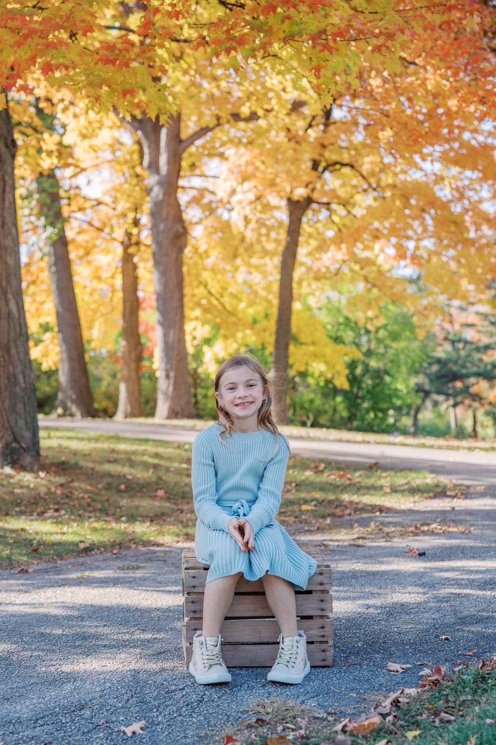 Child in blue sweater sitting on wooden crate surrounded by fall foliage.