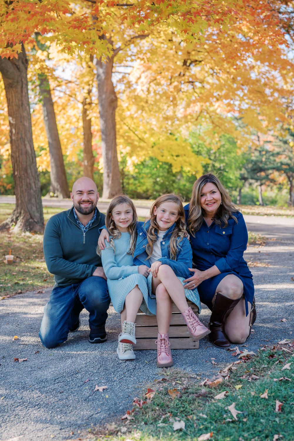 Family group sitting together on path surrounded by golden autumn trees.