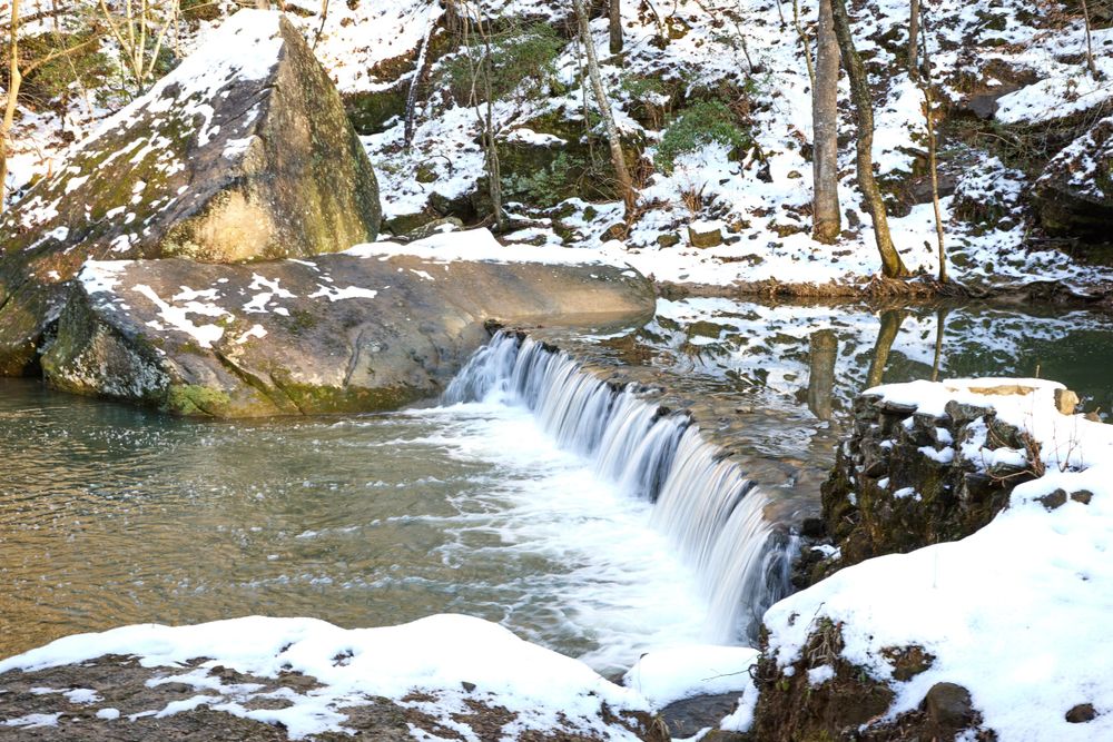Snow blankets the rocks around a gentle waterfall cascade flowing over stone ledges in a winter forest setting.