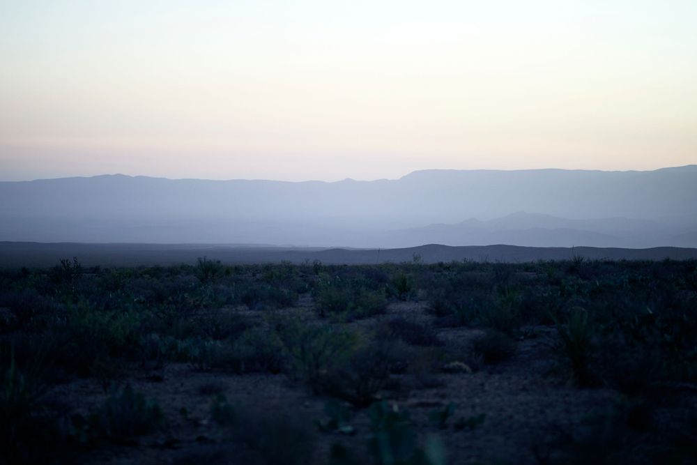 Misty mountain ranges stretch across the horizon at dusk with desert shrubland in the foreground.