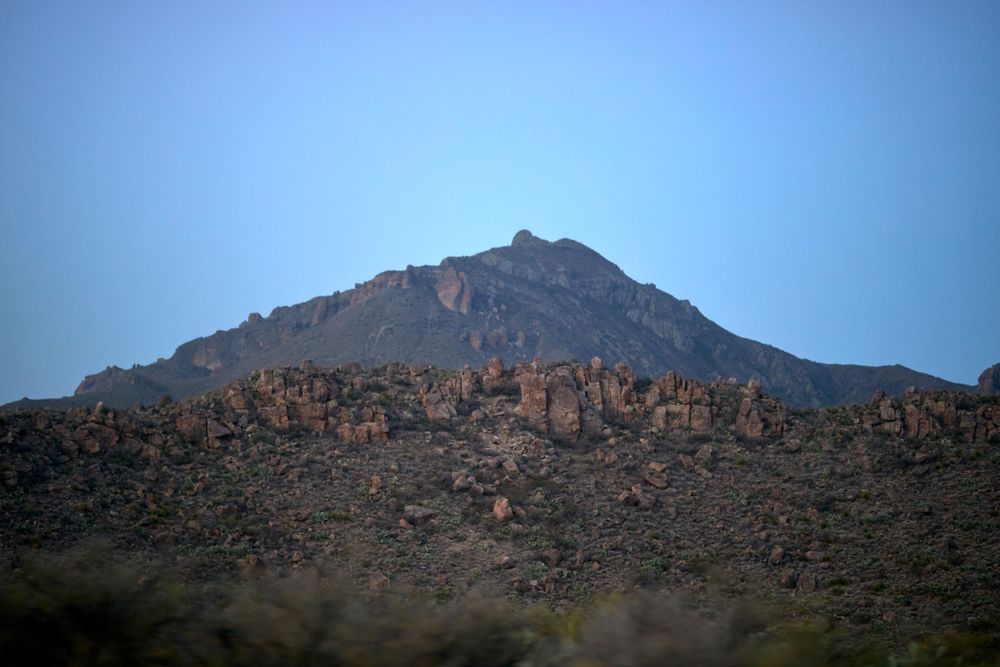 Rocky mountain peak with boulder-strewn slopes stands prominently against clear blue desert sky.