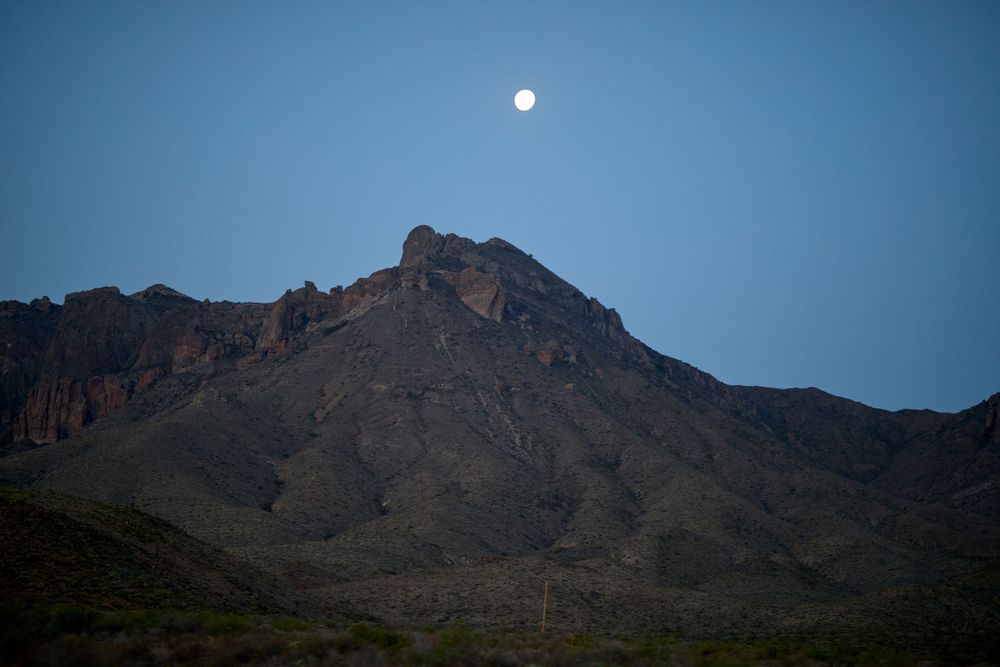 Full moon rises over a rugged mountain peak against a dusky blue sky in the desert landscape.