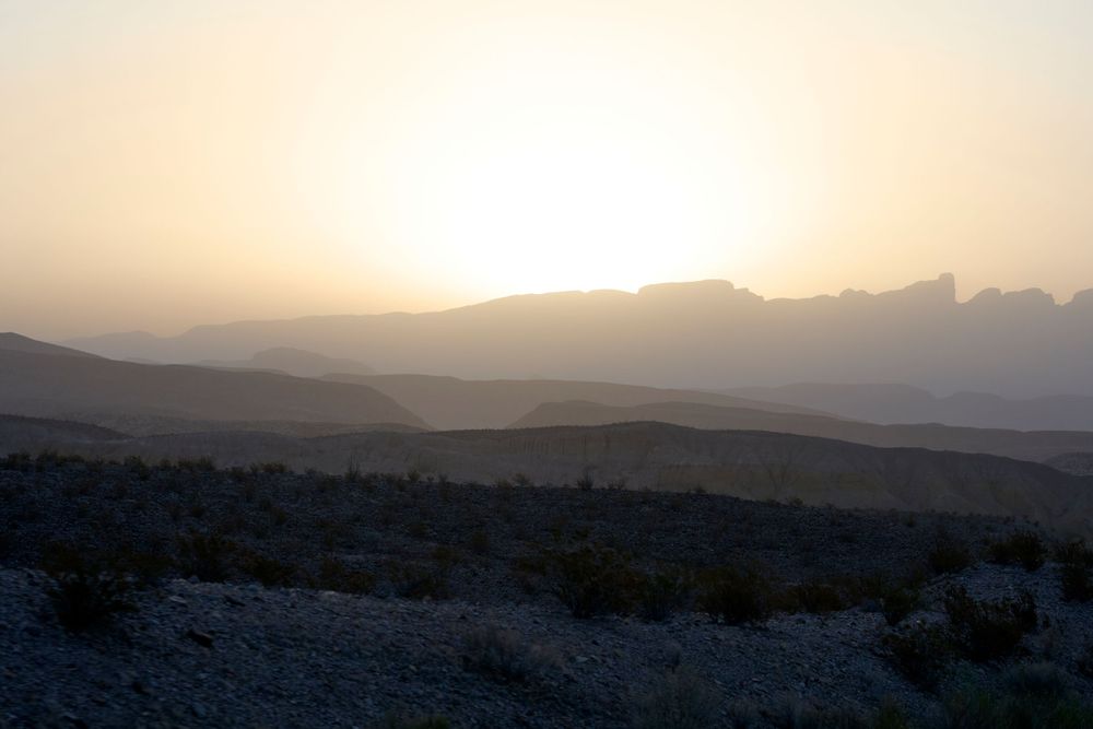 Mountains silhouetted against a warm sunset sky with sun peeking over the horizon in desert landscape.