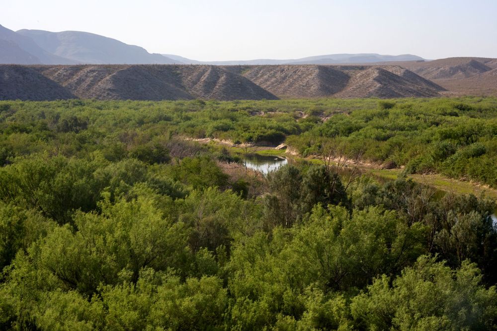 A lush green river valley winds through arid desert mountains in a remote wilderness landscape.