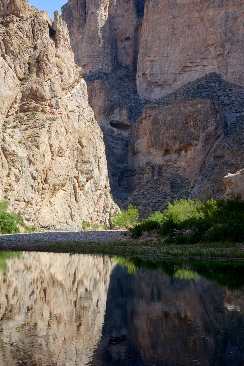 Towering canyon walls reflect in the still waters of a river with green vegetation along the rocky shoreline.