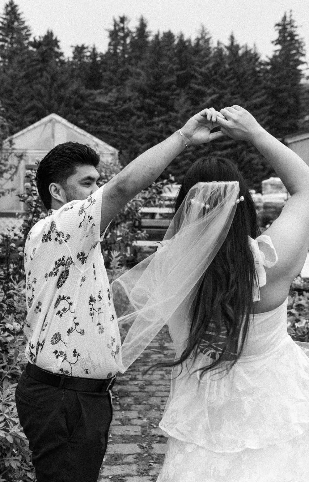 A joyful dancing moment captured in black and white with evergreen trees and a rustic building in the background.