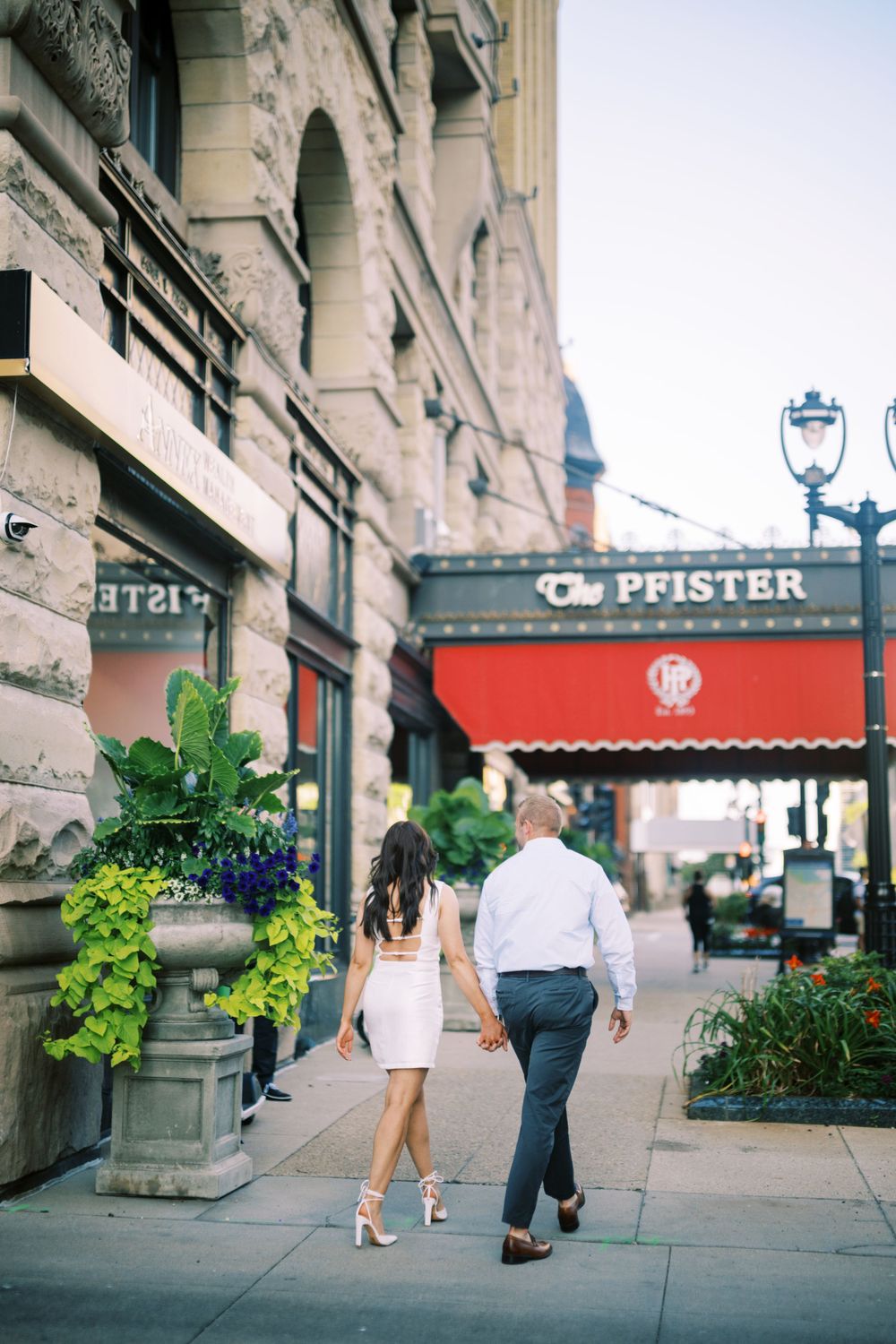 A couple walks together on a city sidewalk past ornate architecture and a Pfister storefront with red awning.