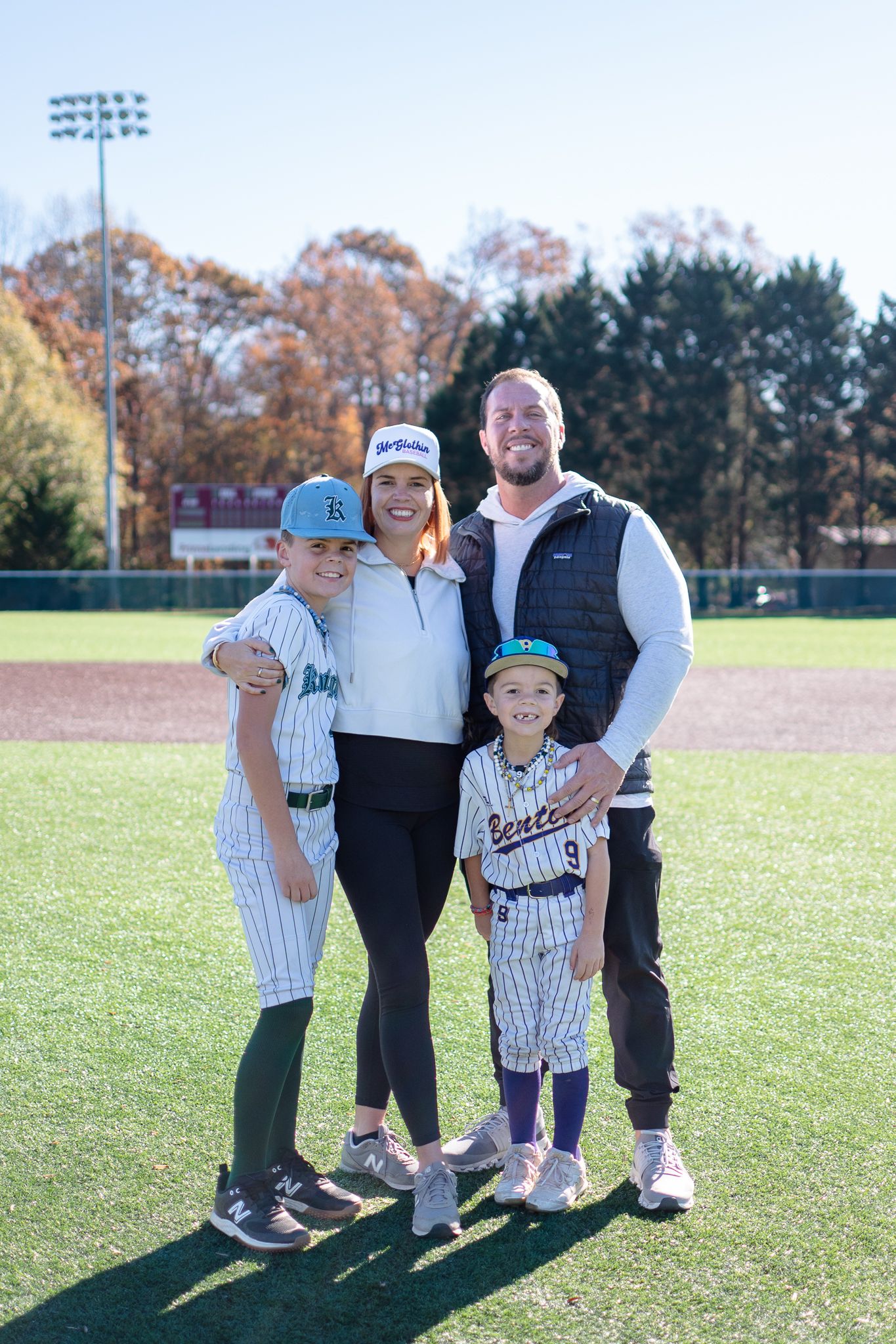 A family stands together on a baseball field with fall trees in the background during a sunny day.
