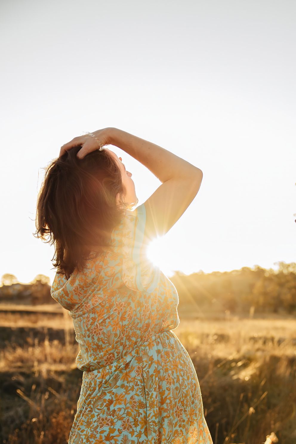 Silhouette of a person in a floral dress standing in a golden field at sunset, backlit by warm sunlight.