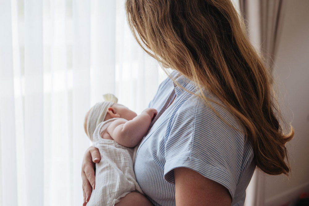 Mother holding newborn baby by window in soft natural light.