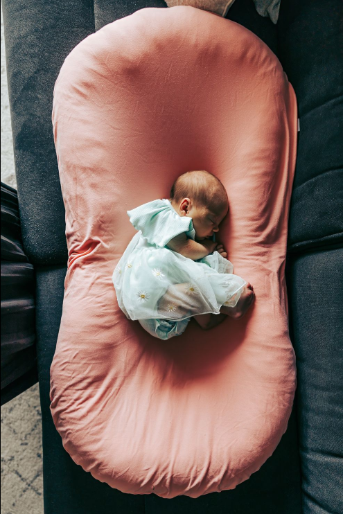 Baby in white onesie sleeping on pink lounger cushion.