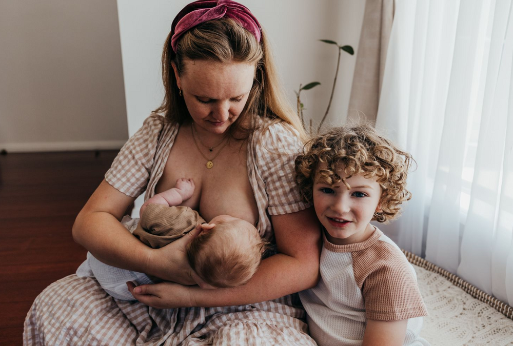Mother nursing baby while toddler sits nearby in cozy indoor setting.