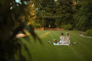 Two figures enjoy an elegant picnic on a blanket in a lush green garden during golden hour.