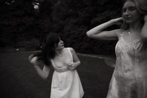 A candid black and white photo captures two friends laughing in white dresses outdoors.