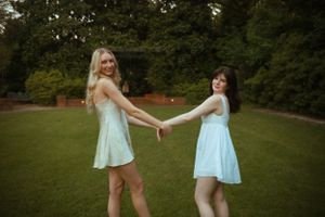 Two friends in light summer dresses hold hands and twirl in a garden.