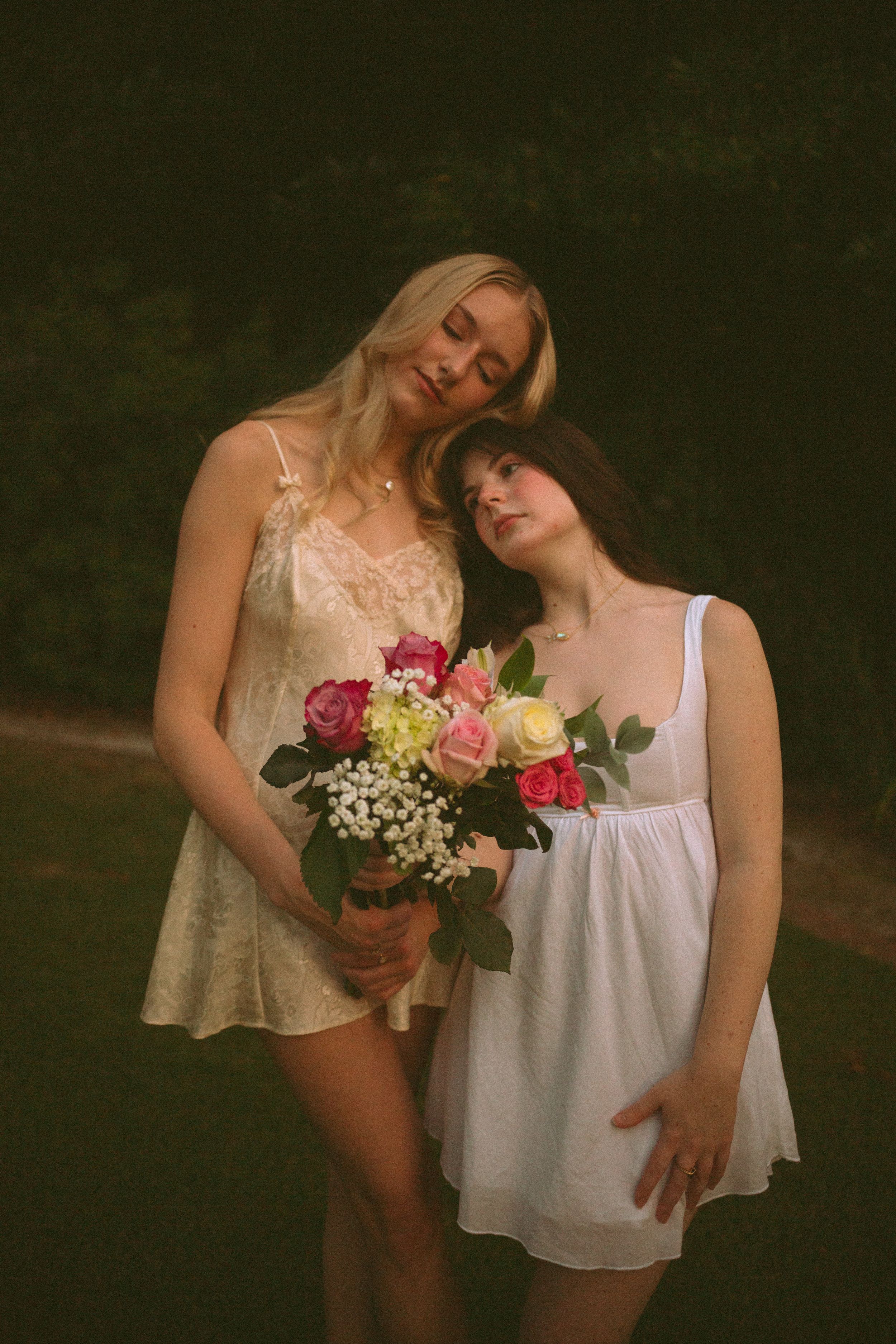 Two friends in white dresses hold a bouquet of pink and yellow roses in a dreamy outdoor portrait.