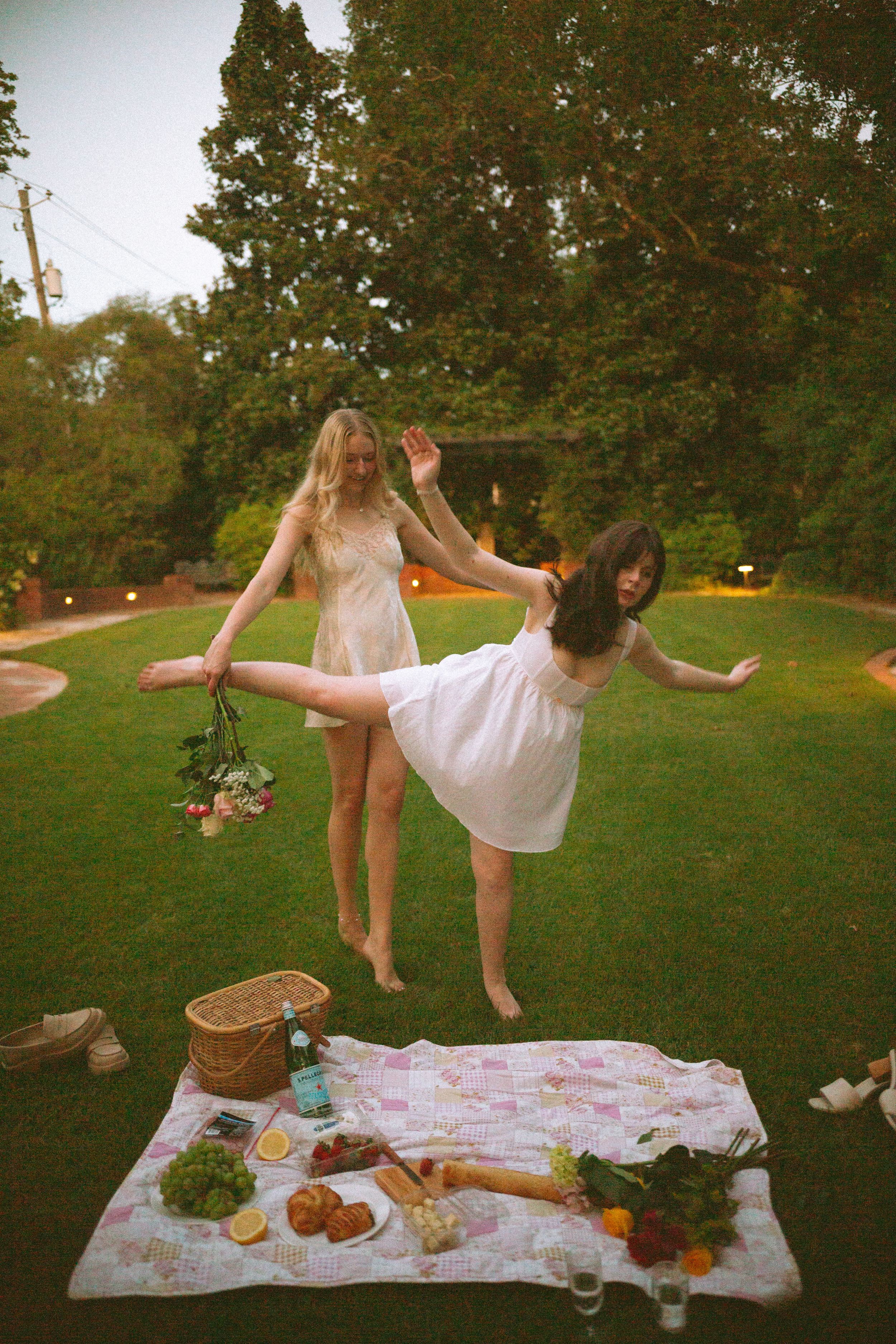 Two friends in white dresses dance and play on a picnic blanket at sunset.