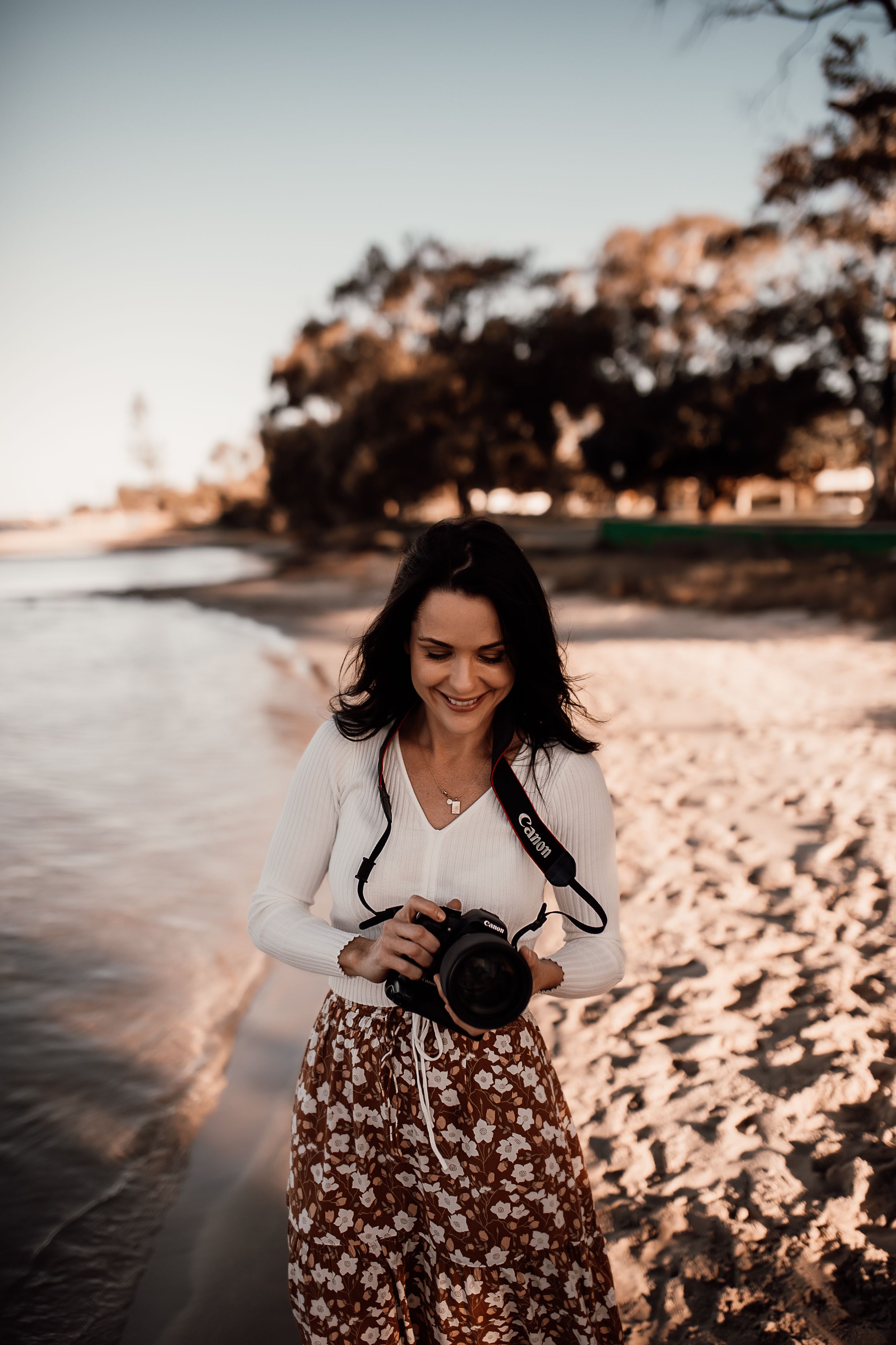 A photographer laughs while holding her camera on a beach at sunset.