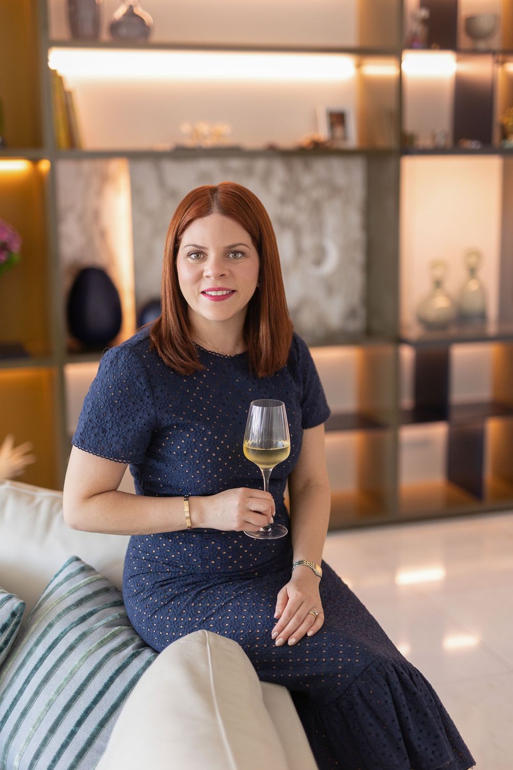 Elegant woman with red hair holding a glass of white wine, seated on a sofa in a stylish, warmly lit interior.