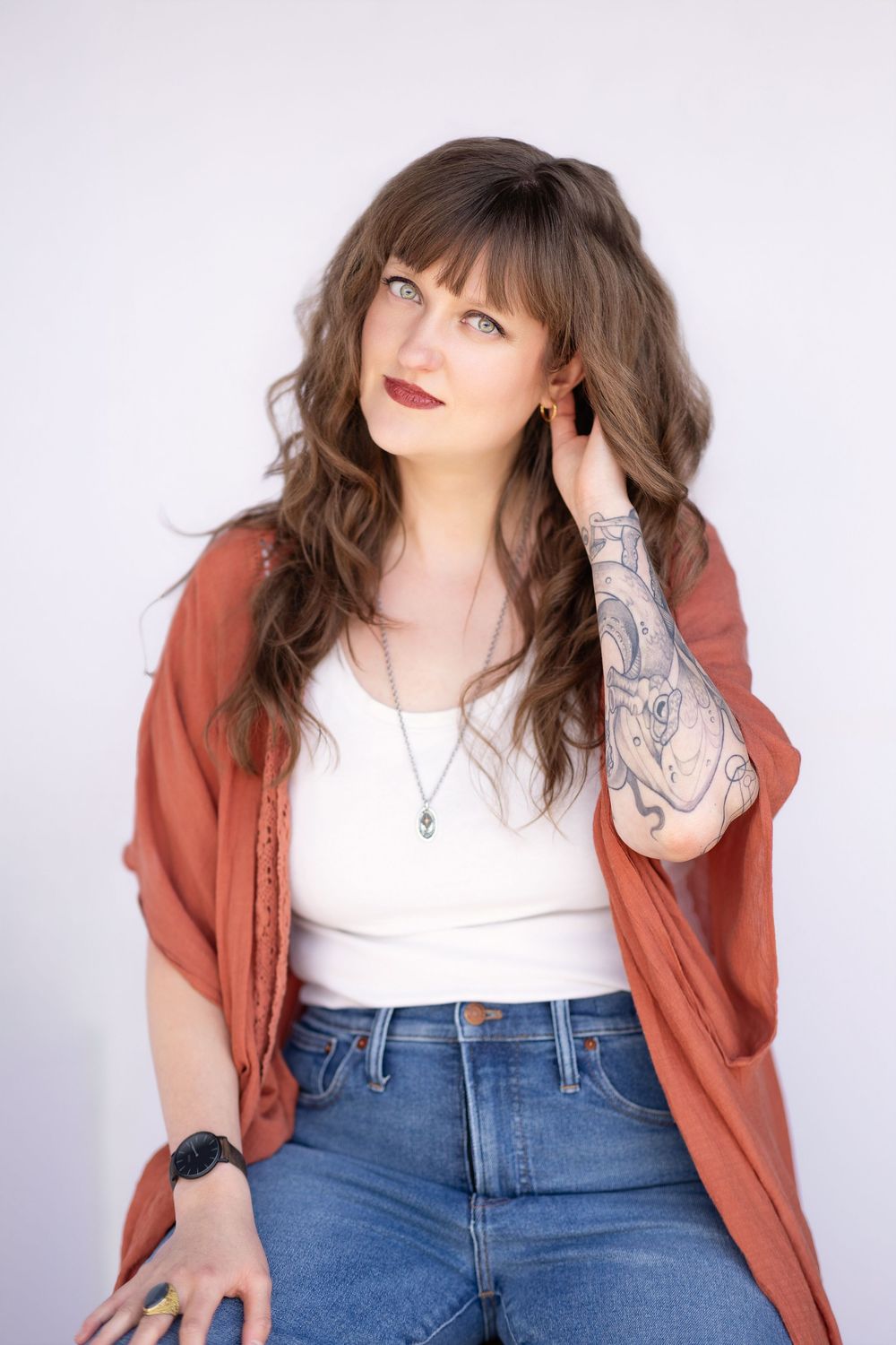 Woman with tattoos in rust cardigan, white top, and jeans poses against white background.