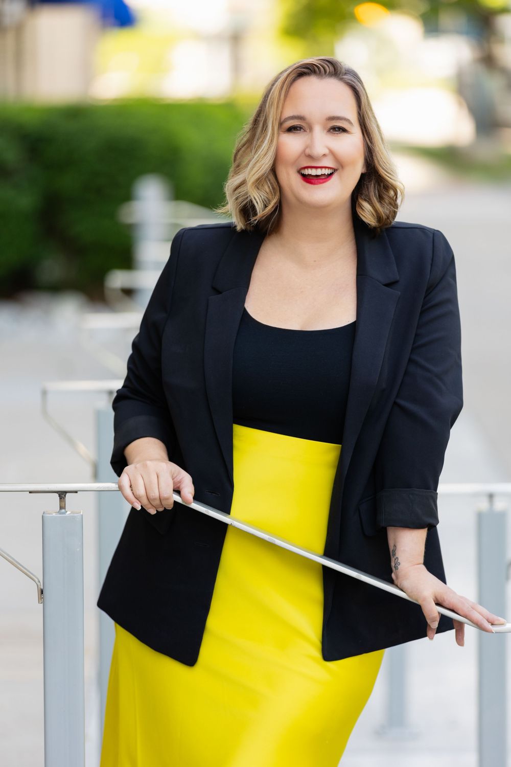 Woman in yellow skirt and black blazer smiling outdoors, leaning on a railing in a professional portrait.