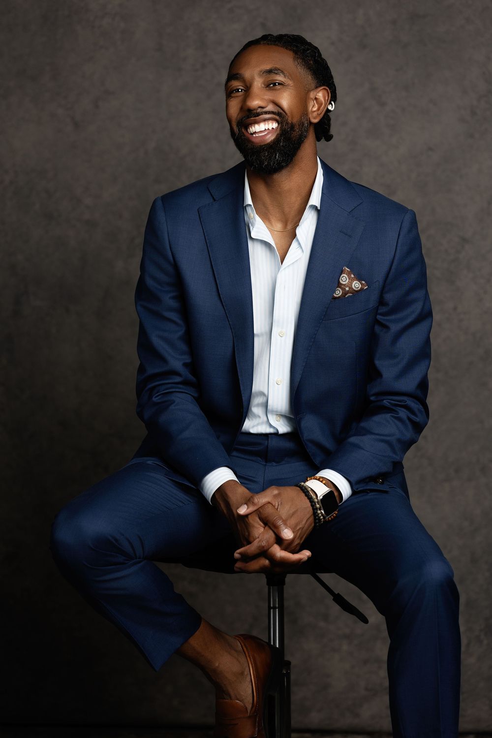 Smiling man in navy blue suit with pocket square sits confidently on stool against gray studio background.