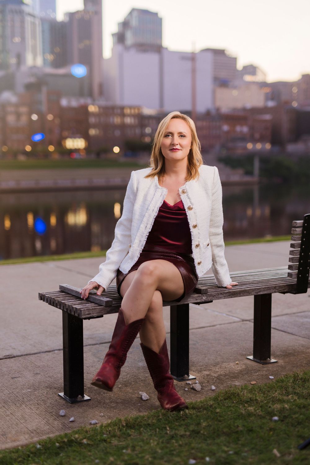Woman in white blazer and burgundy dress sits on bench by urban waterfront at dusk.