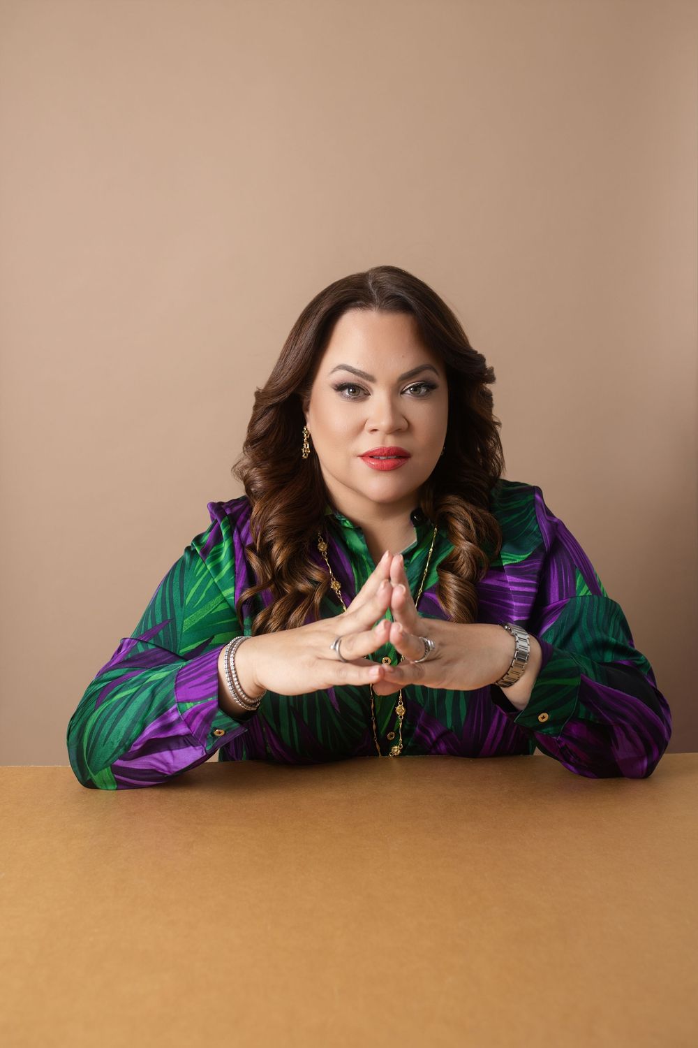 Woman in colorful tropical blouse sits at table with hands pressed together, looking confidently at camera.