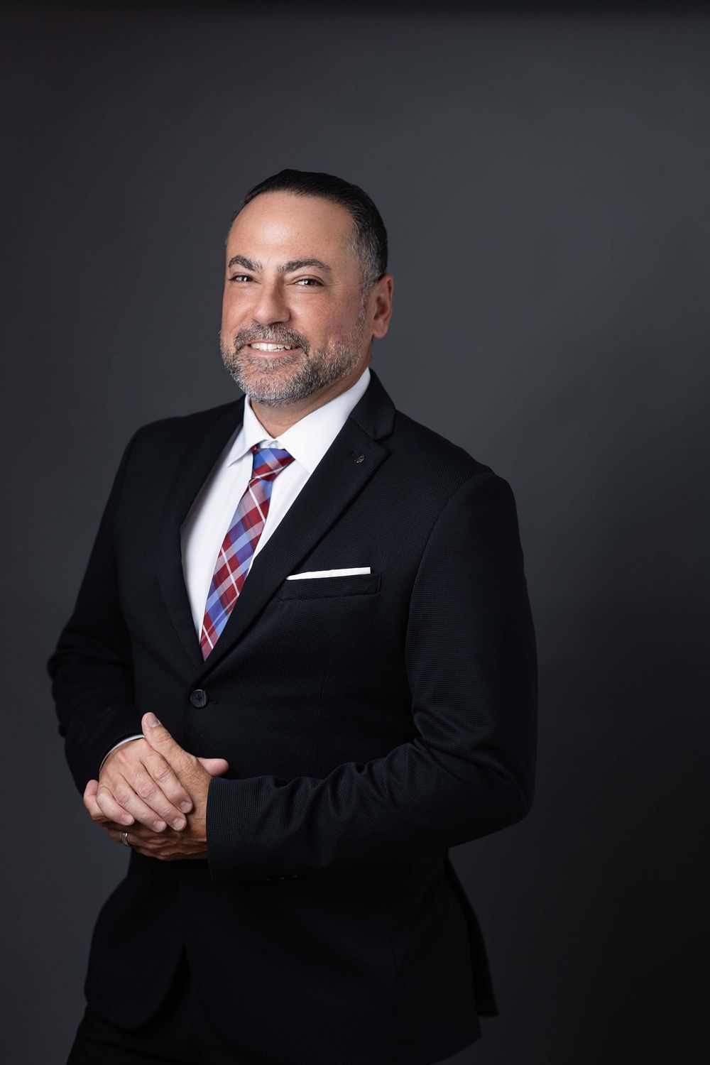 Professional man in black suit and plaid tie smiling confidently against dark gray studio background.
