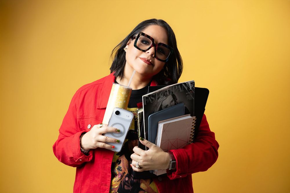 Woman in red jacket and glasses holds notebooks, phone, and mic against yellow background.